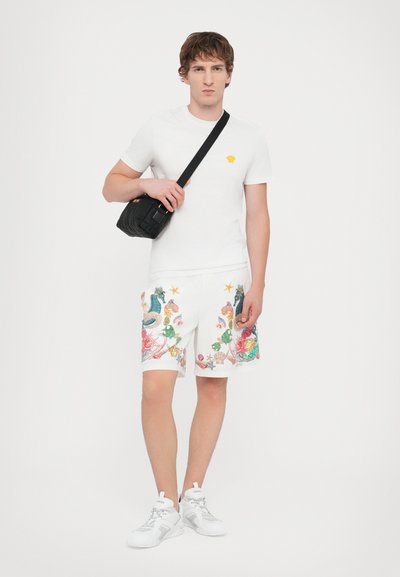 White t-shirt with yellow logo, patterned white shorts featuring colorful marine motifs, and black crossbody bag, worn with white sneakers.