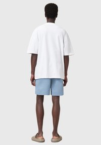 Camiseta de algodón blanca de corte amplio, pantalones cortos azul claro, zapatos sin cordones color tostado. Vista desde atrás, mostrando el corte relajado y el diseño simple.