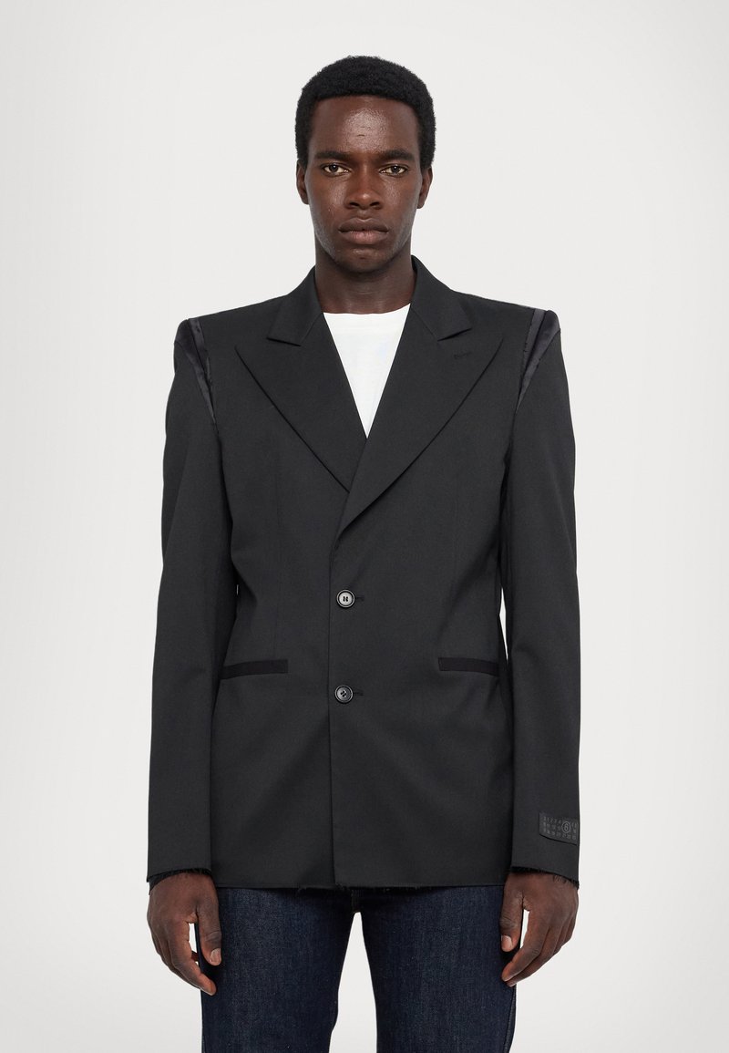 Blazer noir au design ajusté, avec des accents en satin aux épaules, deux boutons sur le devant et des poches inclinées. Porté sur une chemise blanche.