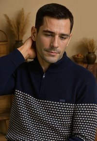 Man in een marineblauw gebreid trui met halve ritssluiting, leunend met zijn hand op zijn nek, zittend binnen met gedroogde planten op de achtergrond.