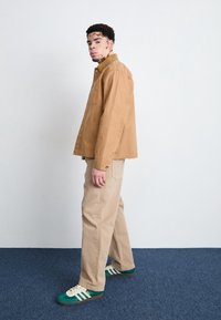 Brun jacka med krage, beige byxor och gröna sneakersdetaljer. Slät textur, avslappnad passform, detaljerad med knappar och fickor.