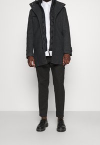 Manteau gris foncé avec une doublure intérieure noire matelassée, doté d'une capuche, de poches à boutons et d'une coupe structurée. Porté par-dessus une chemise blanche et un pantalon foncé.