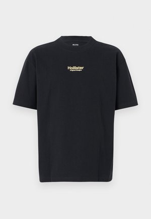 Black cotton t-shirt featuring short sleeves, crew neckline, and front logo in gold text that reads "Hollister Original Designs."