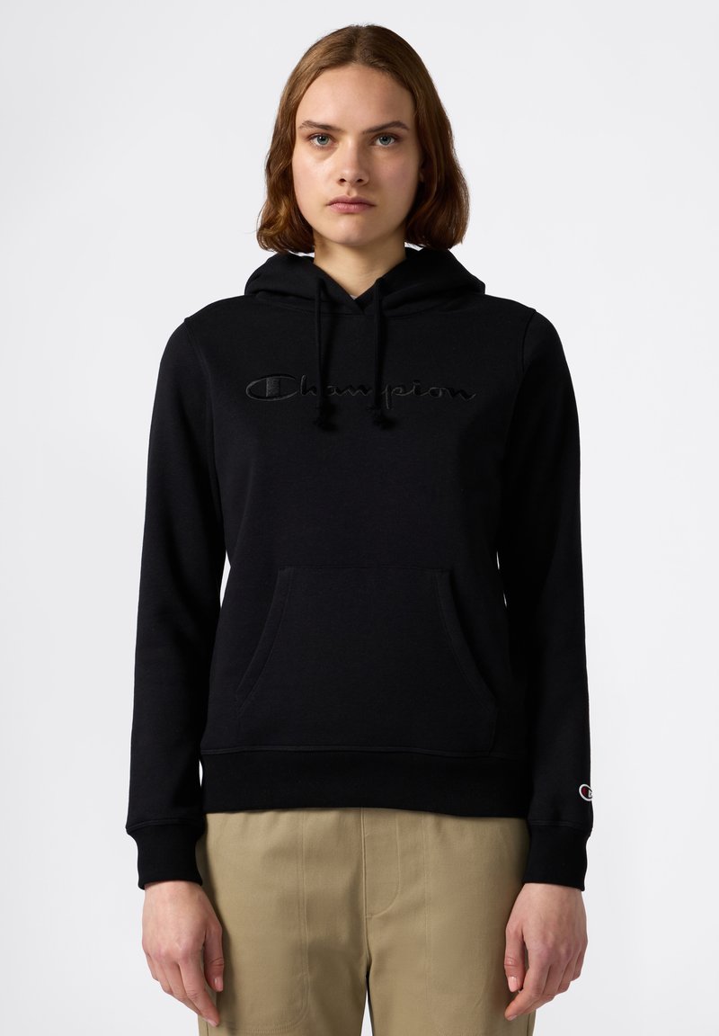 Hoodie in cotone nero con tasca a scomparsa, logo "Champion" ricamato sul petto e lacci sul cappuccio. Indossato con pantaloni beige.