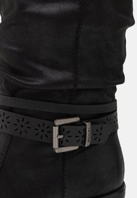 Botte en cuir noir à finition texturée, avec une ceinture décorative découpée à la cheville, agrémentée d'une boucle métallique et d'un accent.