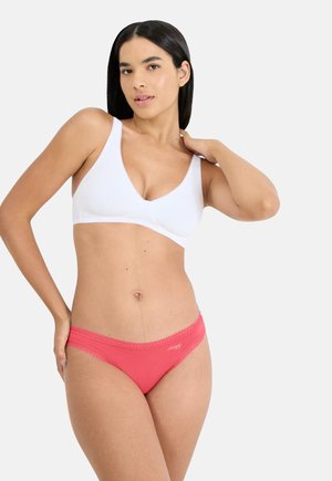 Nő pózol, fehér vezeték nélküli melltartót és korallszínű csipkével díszített alsóneműt visel, egyszerű világos háttér előtt.