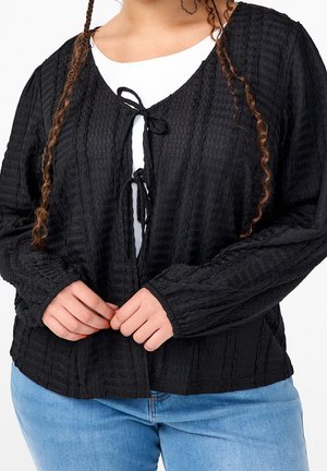 Kobieta ubrana w teksturowany czarny kardigan z wiązaniami z przodu, na białym topie i jasnoniebieskich dżinsach, ręce poprawiają przód kardiganu.