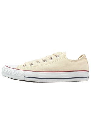 Beiger Sneaker aus Canvas mit weißer Gummisohle, Metallösen und roten und schwarzen Streifen auf der Sohle, von der Seite gezeigt.