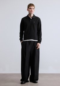 Mustast pikast varrukast polosärk kraega ja rohelise logoga, kombineeritud lahtiste mustade pükstega. Kangas näib olevat sile ning disain on vabaaja stiilis.