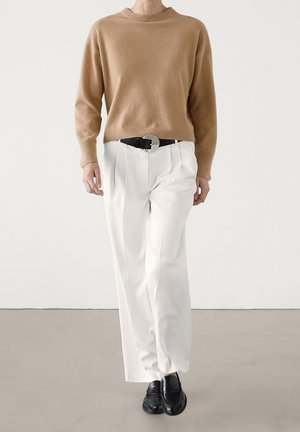 Persona che indossa un maglione color cammello, pantaloni bianchi plissettati con cintura nera e scarpe di pelle nere, in piedi davanti a uno sfondo semplice.