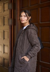 Chaqueta marrón acolchada con capucha, que presenta un cierre frontal y bolsillos laterales, exhibida sobre un fondo de madera. Textura suave.