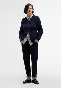 Marinenavy brei cardigan met gestreept patroon, voorrits en kraag, gelaagd over een gestreept shirt, gecombineerd met donkere broek en zwarte schoenen.