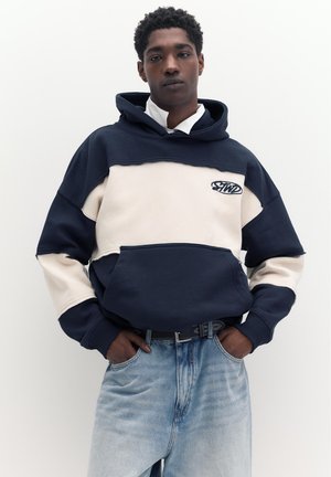 Ung mand iført marineblå og hvid hættetrøje med logo, hænderne i lommerne og lyseblå jeans mod en ensfarvet hvid baggrund.