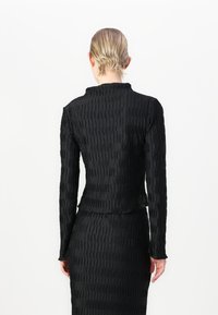 Juodas ilgomis rankovėmis viršus su tekstūruotu, pūstų dizainu. Aukštas apykaklės ir prigludusi siluetas. Lygus audinys, derinamas su atitinkamomis sijoninėmis.