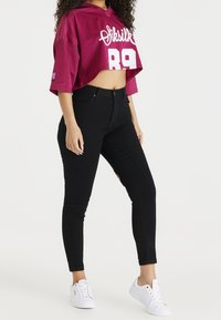 Sudadera burdeos recortada con letras blancas, combinada con vaqueros negros ajustados de talle alto. La modelo lleva zapatillas blancas.