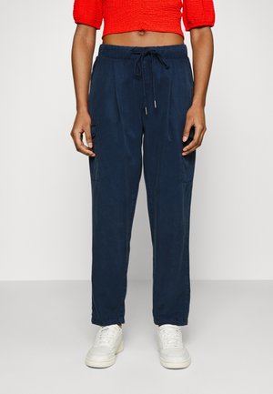 Personne portant un pantalon cargo bleu marine à cordon, des baskets blanches et un top court rouge, debout devant un fond clair uni.