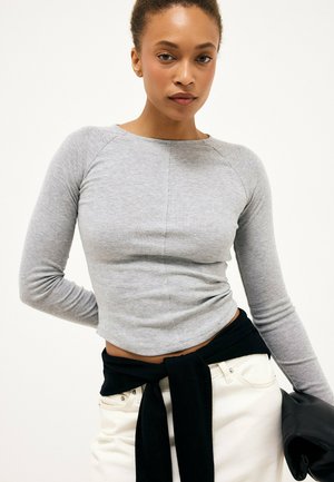 Kvinde iført en tætsiddende grå langærmet top og hvide bukser med en sort sweater bundet omkring livet, der holder en sort jakke.