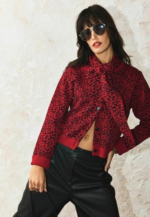 Frau trägt übergroße Sonnenbrille und roten Pullover mit Leopardenmuster und Schluppe, kombiniert mit schwarzer Lederhose, posiert vor einer hellen Wand.