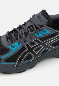 Sapatilha atlética cinza com parte superior em malha, detalhes em preto e azul, apresentando um distinto logotipo da ASICS e sola de borracha texturizada.