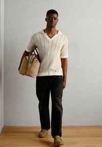 Strickoberteil mit kurzen Ärmeln in Creme mit einem strukturierten Muster, kombiniert mit dunklen Jeans. Das Modell trägt eine Tasche aus braunem Wildleder und trägt braune Schuhe.
