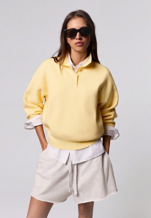 Femme portant des lunettes de soleil noires, un pull jaune sur une chemise blanche et un short beige clair, les mains dans les poches, sur un fond uni.