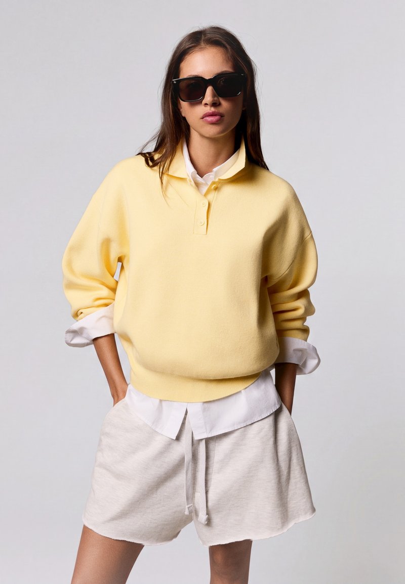 Femme portant des lunettes de soleil noires, un pull jaune sur une chemise blanche et un short beige clair, les mains dans les poches, sur un fond uni.