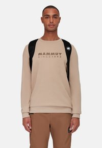 Beige sweatshirt met "MAMMUT SINCE 1862" in bruin gedrukt. Heeft een ronde halslijn en elastische manchetten, gedragen met bruine broek en een zwarte rugzak.