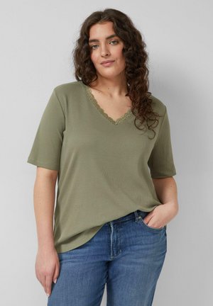 Kobieta o kręconych włosach, ubrana w oliwkowozielony top z dekoltem w serek i niebieskie dżinsy, stoi z jedną ręką w kieszeni na jednolitym tle.