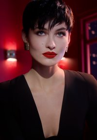 Modell mit einer eleganten, kurzen schwarzen Frisur, tragend kräftigen roten Lippenstift, ein tiefes schwarzes Outfit und goldene Creolen, vor einem roten Hintergrund.