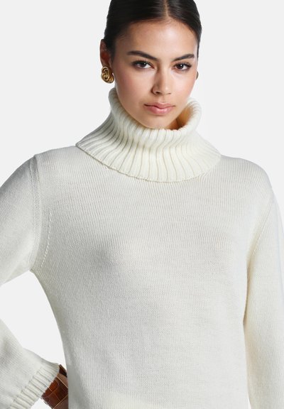 Femme aux cheveux foncés portant un pull à col roulé côtelé blanc et des boucles d'oreilles spirales dorées, posant avec la main sur la hanche devant un fond uni.