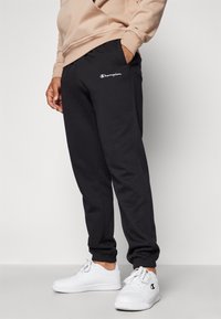 Schwarze Fleece-Sweatpants mit elastischen Bündchen, seitlichen Taschen und gesticktem Champion-Logo in Weiß und Rot am linken Oberschenkel.
