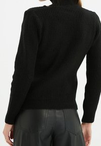 Schwarzer gerippter Strickpullover mit hohem Kragen und figurbetontem Schnitt. Der Stoff hat eine strukturierte Optik und wird mit hoch taillierten Lederhosen kombiniert.