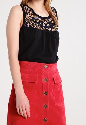 Frau trägt ein ärmelloses schwarzes Oberteil mit bunter Stickerei und einen hoch taillierten roten Rock mit Knopfleiste und großen Taschen.