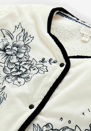 White fleece jacket with black floral embroidery. Features a black trim and snap closures. Soft texture and detailed floral design.
