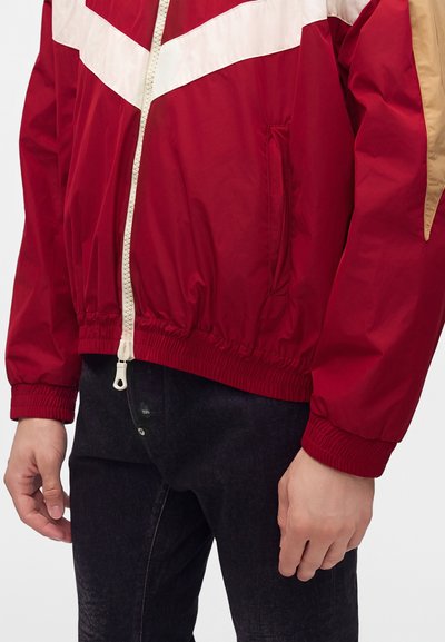 Veste zippée rouge et beige avec poignets et ourlet élastiques portée avec un jean noir, montrant le torse et les bras sans tête visible.