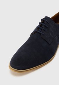 Chaussure en daim bleu marine à bout rond, dotée d'un système de laçage à cinq œillets et d'une semelle en caoutchouc brun clair. Texture lisse.