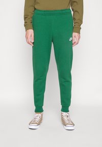 Green sweatpants with elastic waistband and cuffs. White Nike logo on left thigh. Features a smooth texture and tapered design.