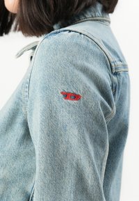 Oseba v svetlom modrem usnjenem jopiču z majhnim rdečim vezenim logotipom na zgornjem delu roke, prikazana s strani in izrameni.