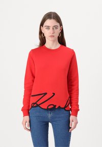 Červená mikina s dlhými rukávmi, ktorá v spodnej časti zobrazuje výrazné čierne logo "Karl". Je vyrobená z hladkej látky a má okrúhly výstrih.