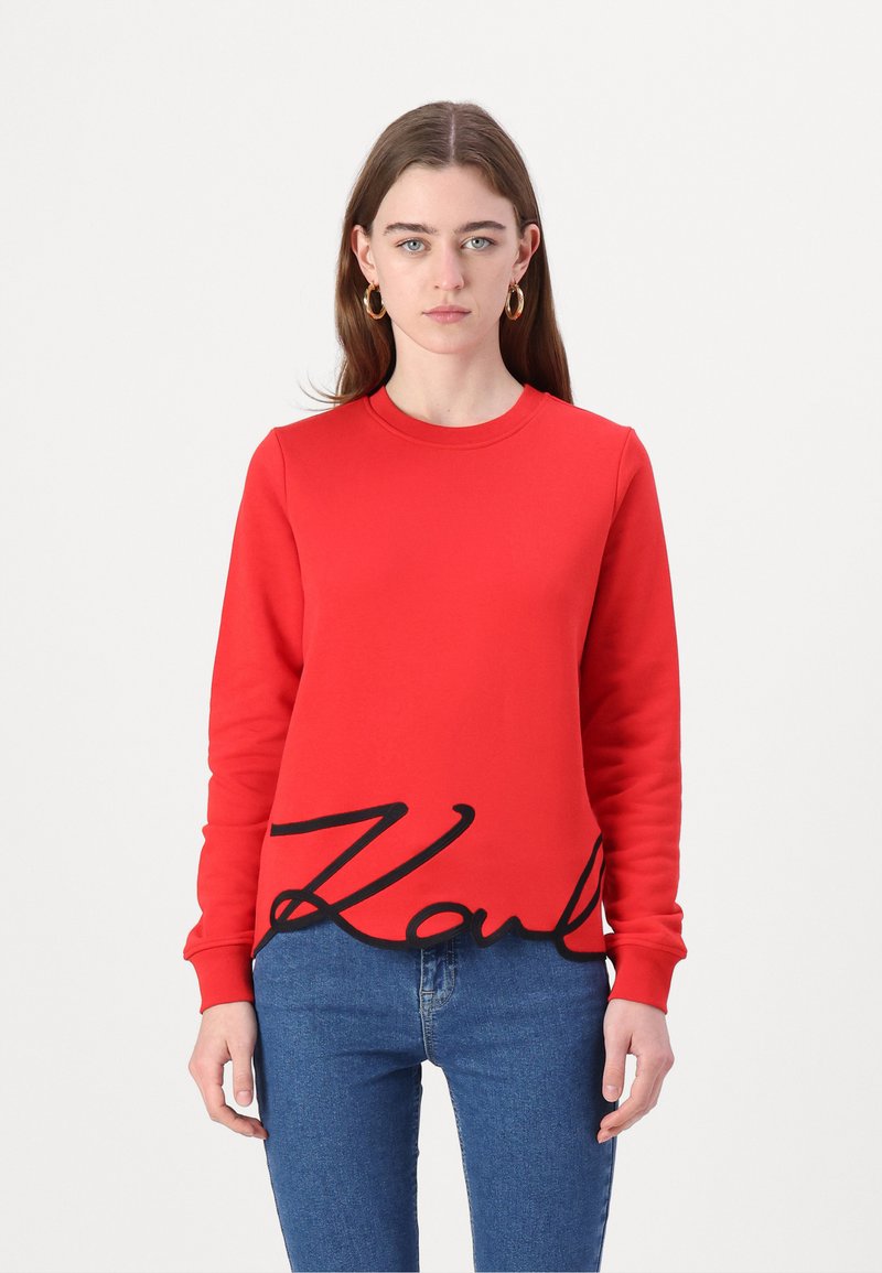 Červená mikina s dlhými rukávmi, ktorá v spodnej časti zobrazuje výrazné čierne logo "Karl". Je vyrobená z hladkej látky a má okrúhly výstrih.