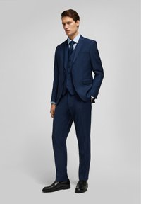 Costume bleu marine avec gilet assorti, coupe ajustée, revers crantés et trois boutons. Une paire de chaussures de ville noires brillantes complète le look.