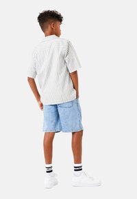 Camisa de rayas de manga corta en blanco y gris claro, combinada con pantalones cortos de mezclilla azul claro y zapatillas blancas, vista desde la parte de atrás.