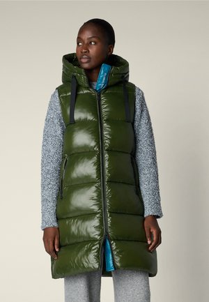 Donna nera che indossa un lungo gilet imbottito verde oliva lucido con cappuccio sopra un maglione e pantaloni grigi strutturati, che guarda di lato.