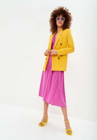 Gelber Blazer über einem rosa Kleid mit einer Twist-Design am Ausschnitt. Kombiniert mit gelben Absatzsandalen und hellvioletten Sonnenbrillen.