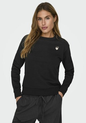 Frau mit langen hellbraunen Haaren, die einen schwarzen Pullover mit „LOVE“ und Herzmotiv auf der Brust sowie dunkle Hose mit Kordelzug trägt, Hände in den Taschen.