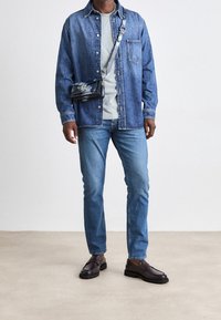 Džínová košile, světle šedé tričko, modré džíny, tmavě hnědé boty, kabelka přes rameno s vzorovaným designem. Ležérní outfit s volným střihem.