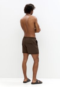 Braune Schwimmshorts mit einer Gesäßtasche, elastischem Bund und seitlichen Schlitzen. Getragen mit schwarzen Sandalen. Das Modell steht mit verschränkten Armen, den Rücken zur Kamera.