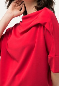 Röd topp med draperad halsringning, korta ärmar och slät textur. Tyget verkar lätt och har en avslappnad design.
