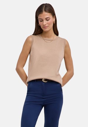 Donna con lunghi capelli castani che indossa un top beige senza maniche, gioielli d'oro e pantaloni blu navy con cintura nera, in piedi con le mani dietro la schiena.