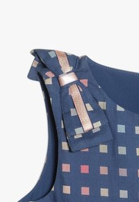 Tmavě modrá látka s mřížkovým vzorem pastelových čtverců. Obsahuje mašli s kovovými akcenty na ramenním popruhu.